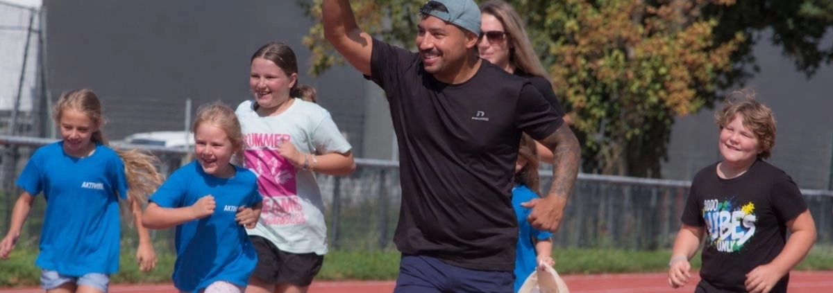 Ein erwachsener Mann läuft zusammen mit mehreren lachenden Kindern auf einer Outdoor-Laufbahn; einige winken und tragen Sportkleidung.