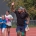 Ein erwachsener Mann läuft zusammen mit mehreren lachenden Kindern auf einer Outdoor-Laufbahn; einige winken und tragen Sportkleidung.